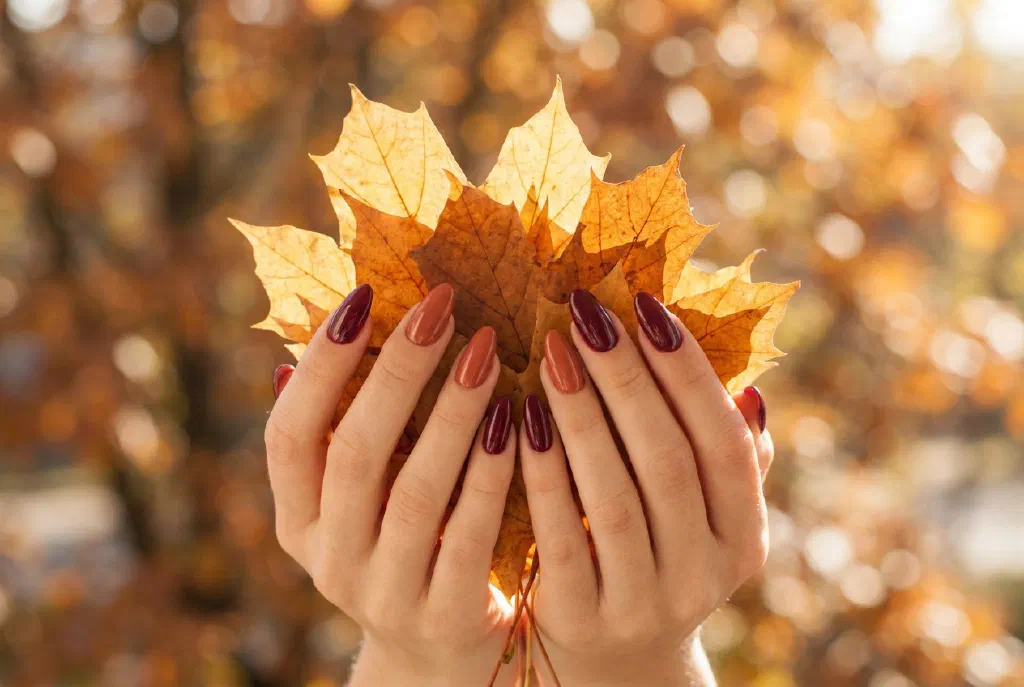 Hands with warm autumn nails in burgundy and terracotta tones against fall foliage
